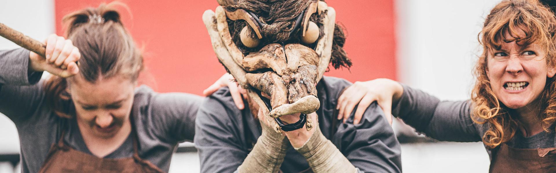 a street theatre show with the performers wearing scary animal masks in front of a red Eisteddfod sign