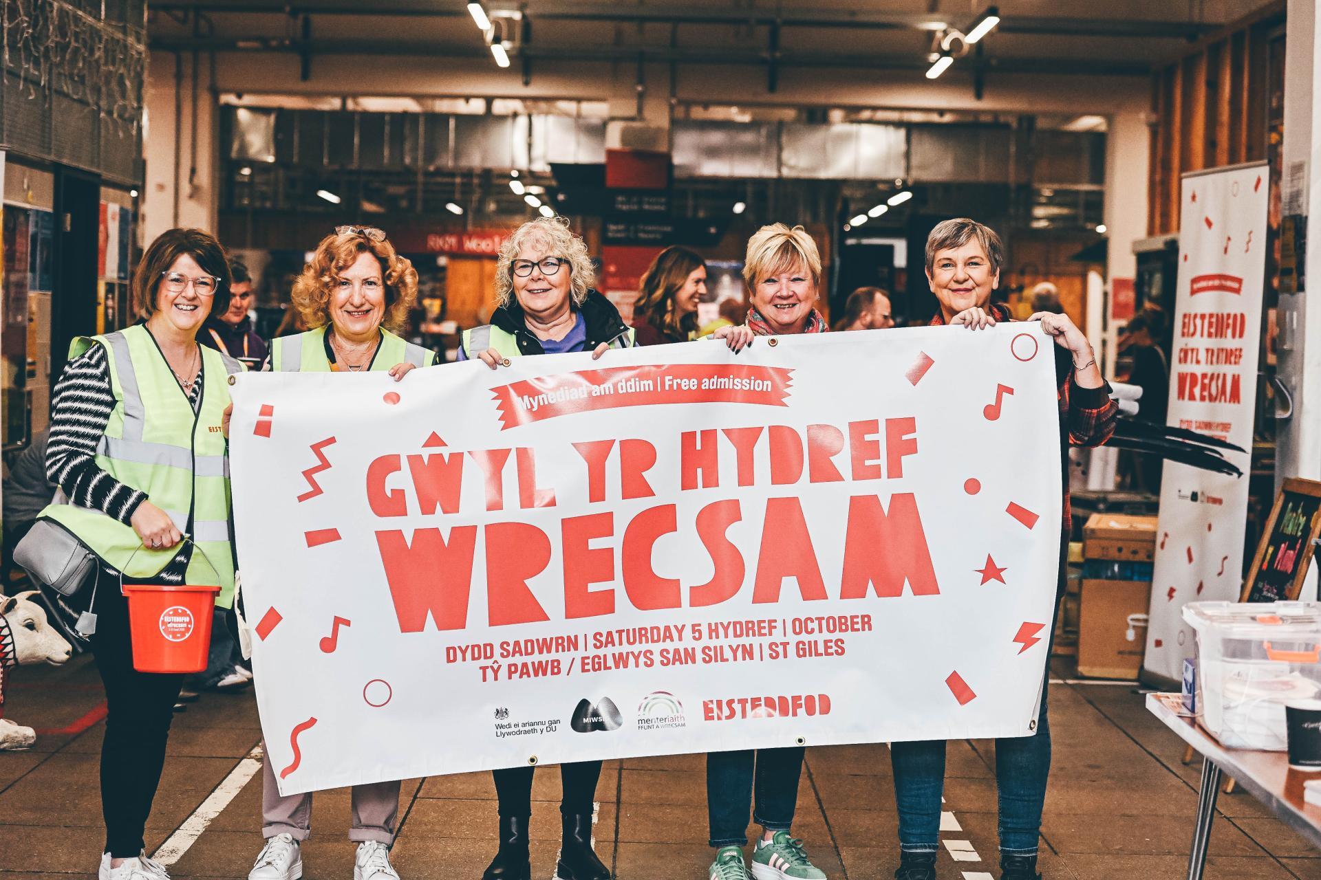 5 women standing behind a white banner with red writing wearing hi-vis jackets against a busy background inside a building