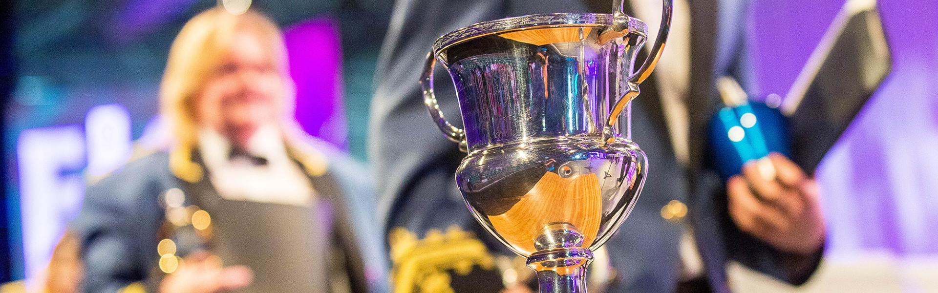 Silver trophy on display on a table on the stage with members of a brass band standing behind.  Purple background