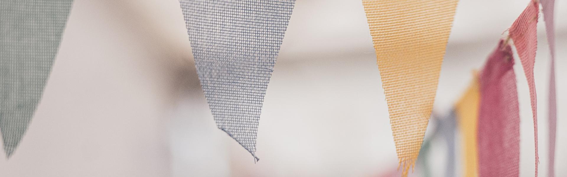 Close-up of colorful fabric bunting flags hanging on a string, with a blurred background showing more bunting flags in various colours.