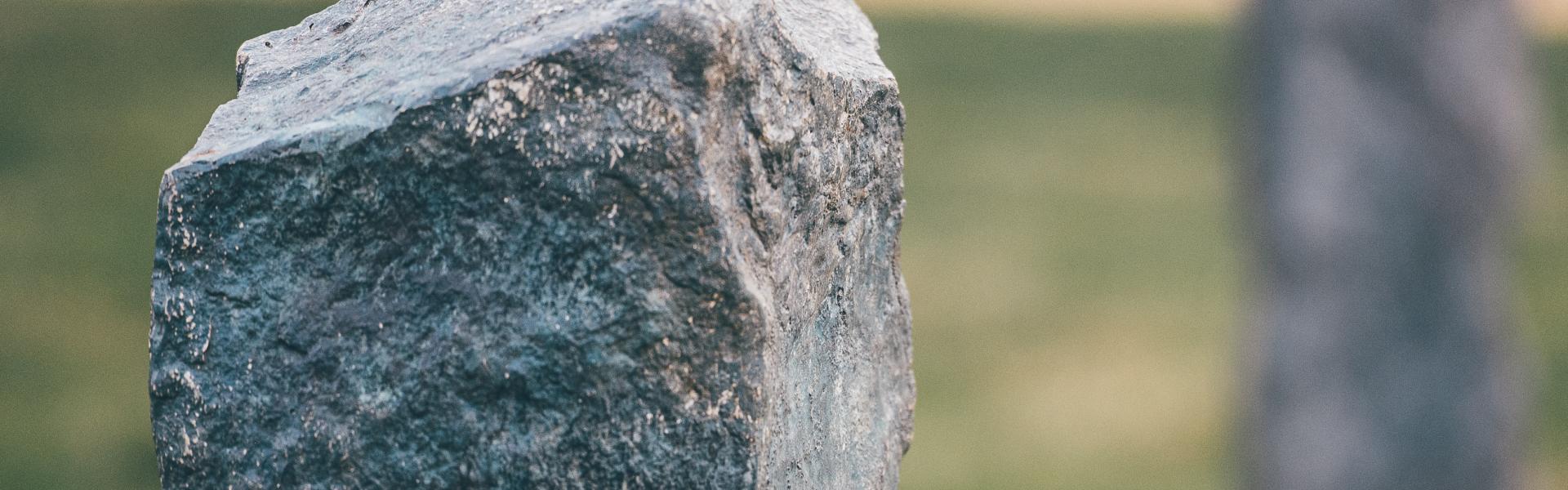 Close-up of a weathered standing stone in a grassy outdoor setting, with a second stone visible in the background. A wooden fence is faintly visible behind them