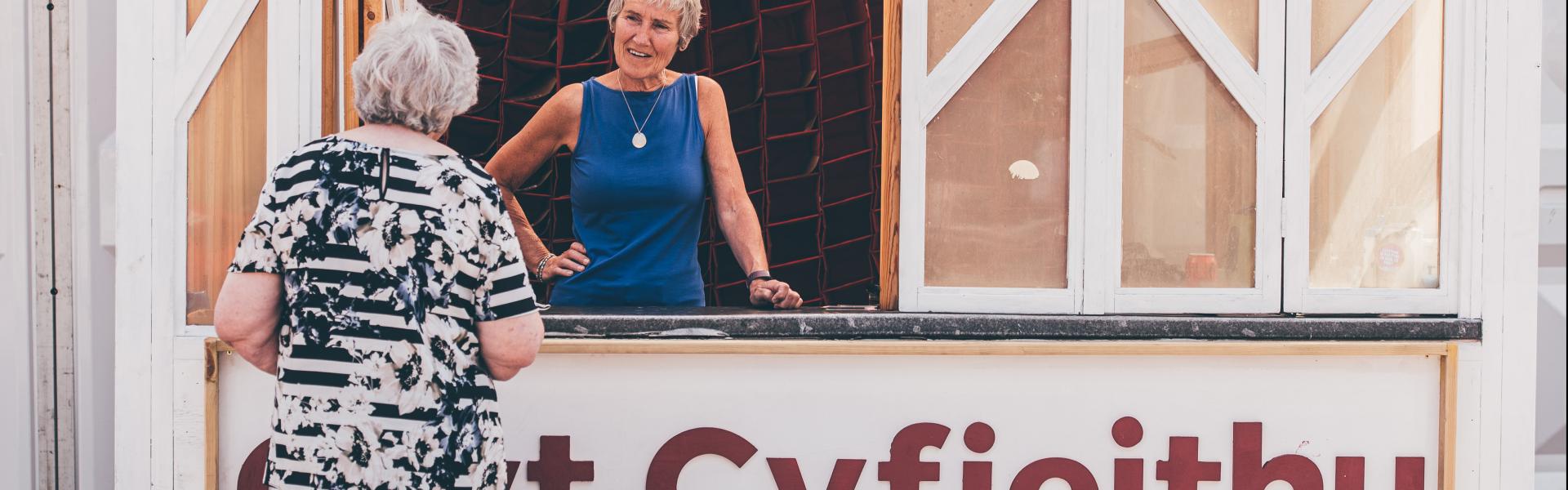 Two people interacting at a wooden booth labeled 'Cwt Cyfieithu Translation Hut'; one person stands inside behind a glass window, and the other stands outside facing the booth