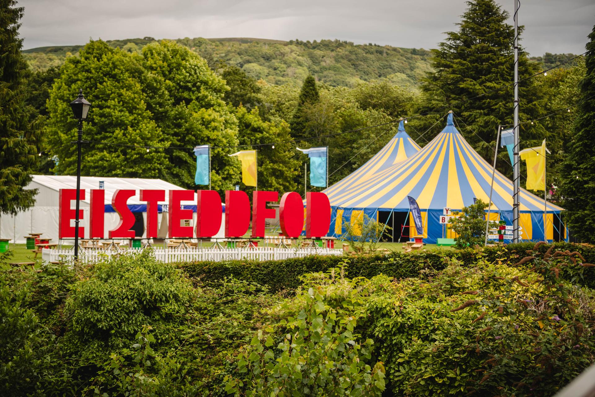 Colorful festival scene with a large blue and yellow striped tent and a red 'EISTEDDFOD' sign in front, surrounded by green trees, smaller tents, and flags under a cloudy sky