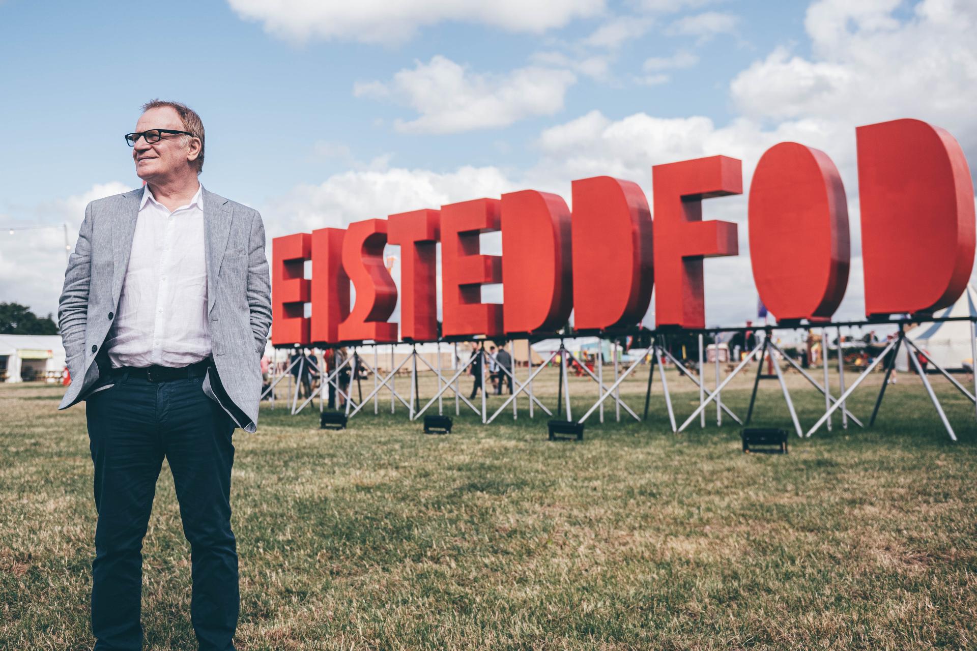 Man in grey blazer, white shirt, and dark pants standing on grass in front of a large red 'EISTEDDFOD' sign with a partly cloudy sky and tents in the background
