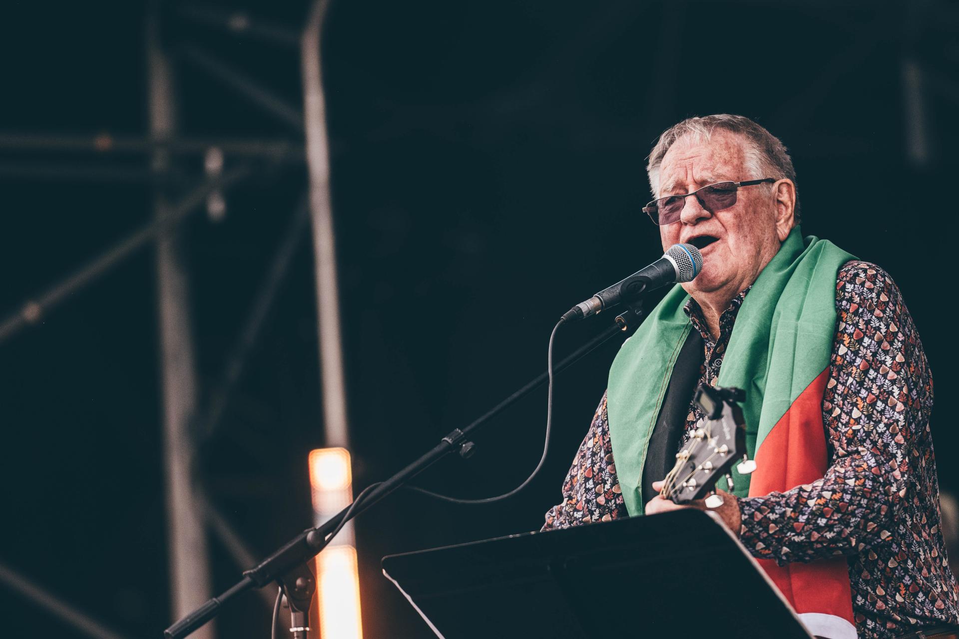 Person on stage holding a guitar and singing into a microphone, wearing a patterned shirt and draped in a Palestinian flag, with stage equipment and lighting in the background
