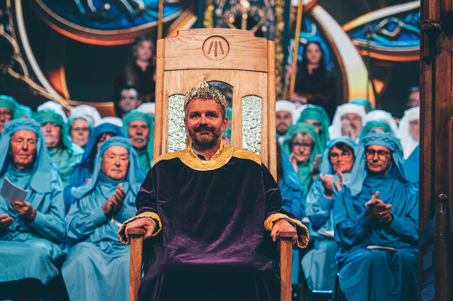 Person in a dark purple and gold robe seated on an ornate wooden throne, with individuals in blue robes behind them, set against a richly decorated background.