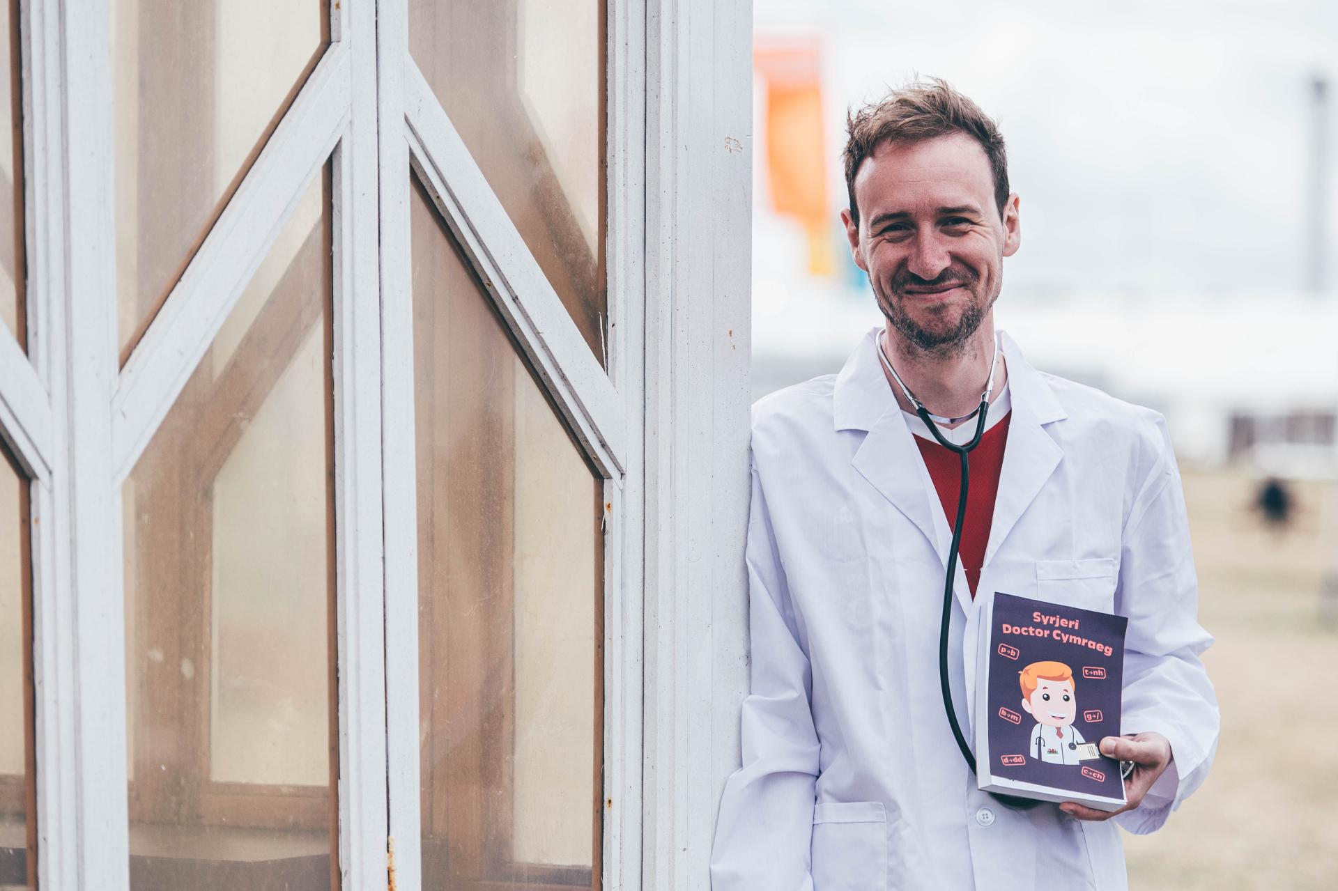 Person in a white lab coat holding a prescription pad titled 'Syrjeri Doctor Cymraeg',and a building with glass windows in the background