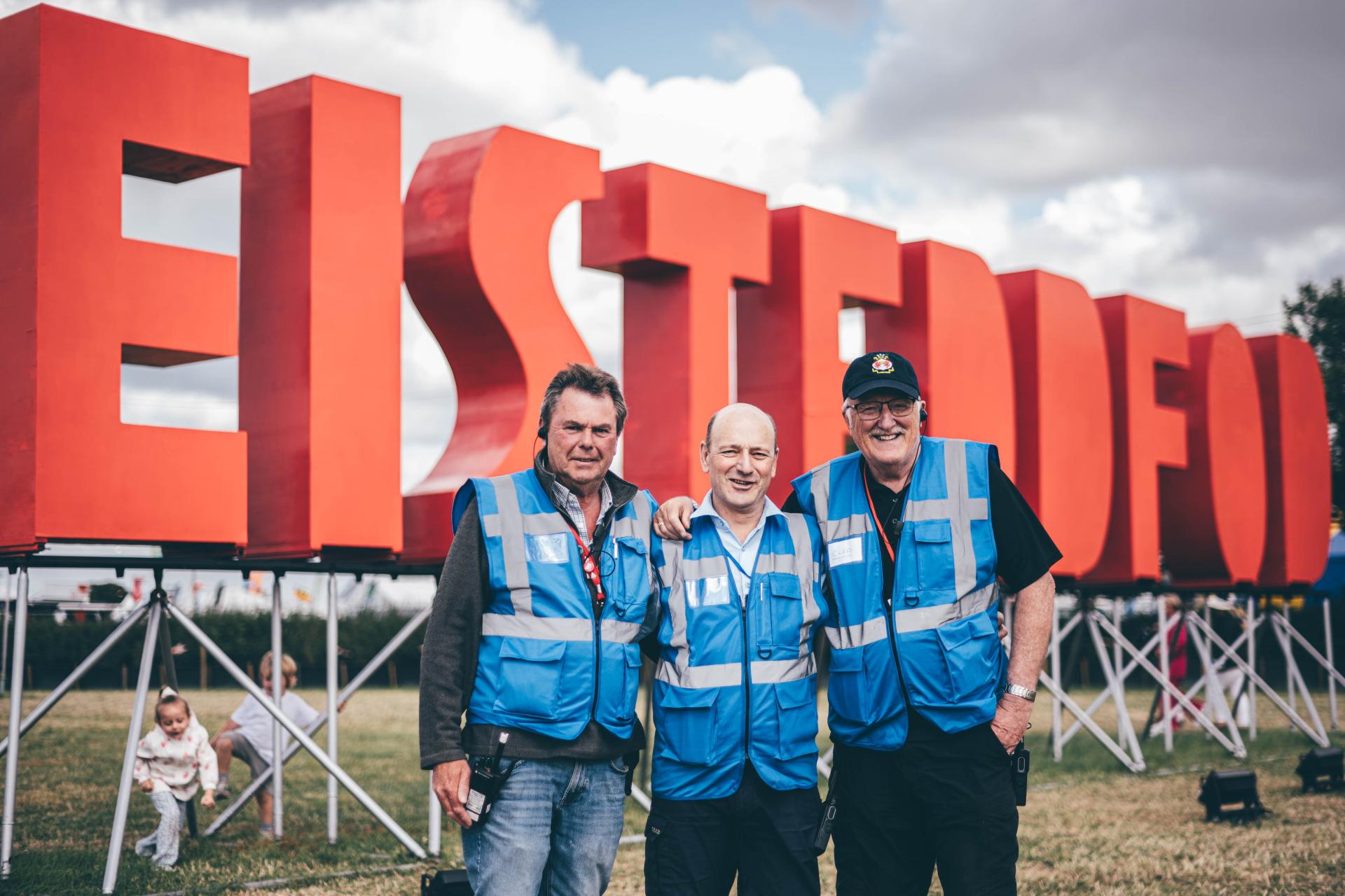 Three volunteers in blue reflective vests standing in front of large red 'EISTEDDFOD' letters, with children playing on the grass in the background under a cloudy sky