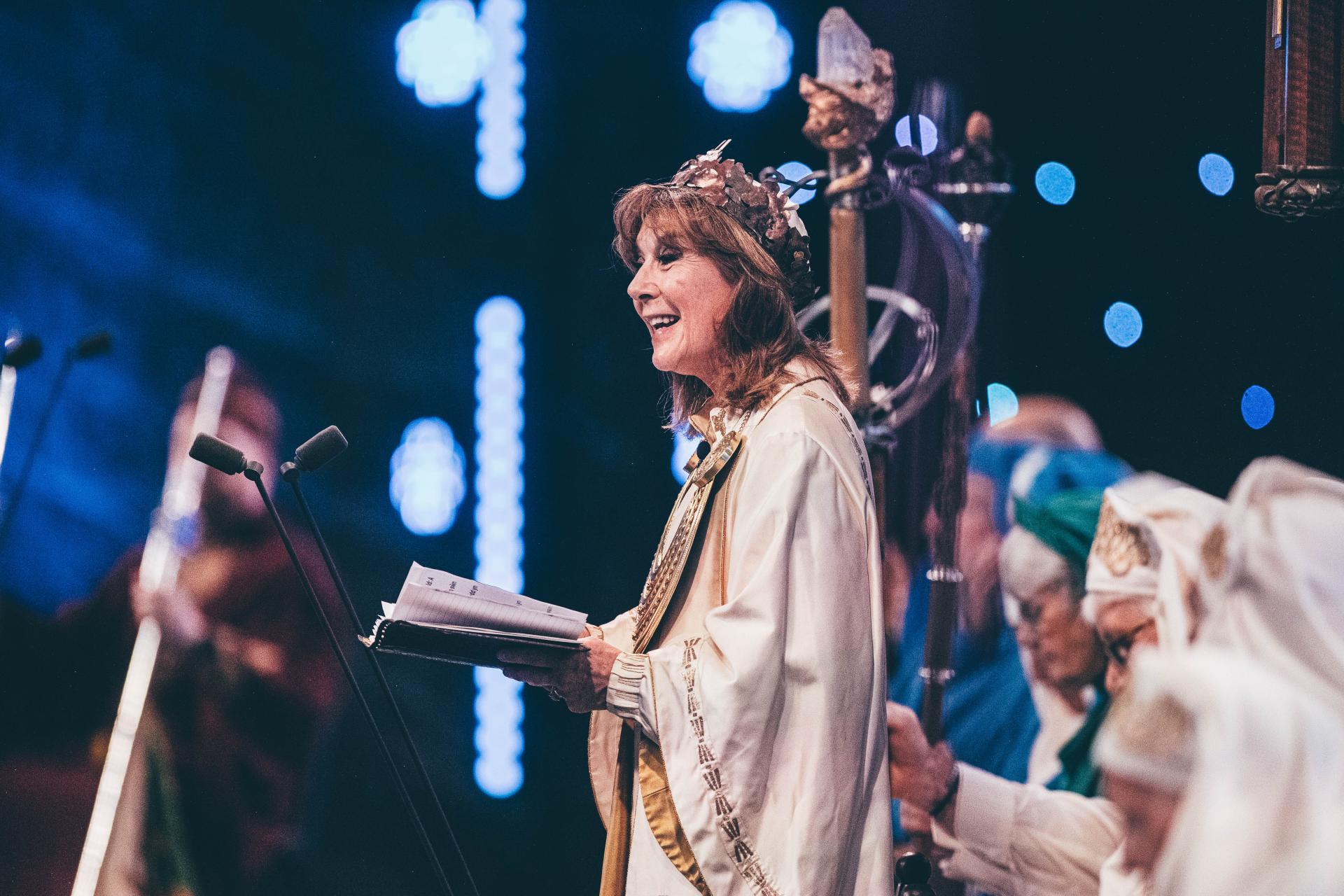 Ceremony with individuals in ceremonial robes, central figure holding and reading from an open book, with bright lights and similarly dressed participants in the background
