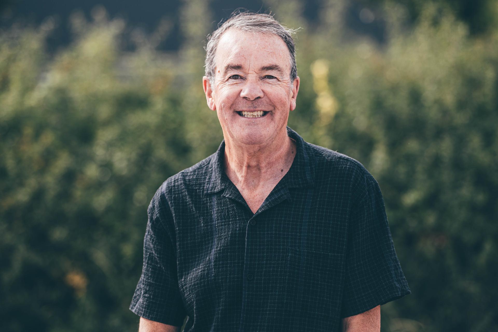 Person in a dark short-sleeved shirt standing outdoors against a green, leafy background 