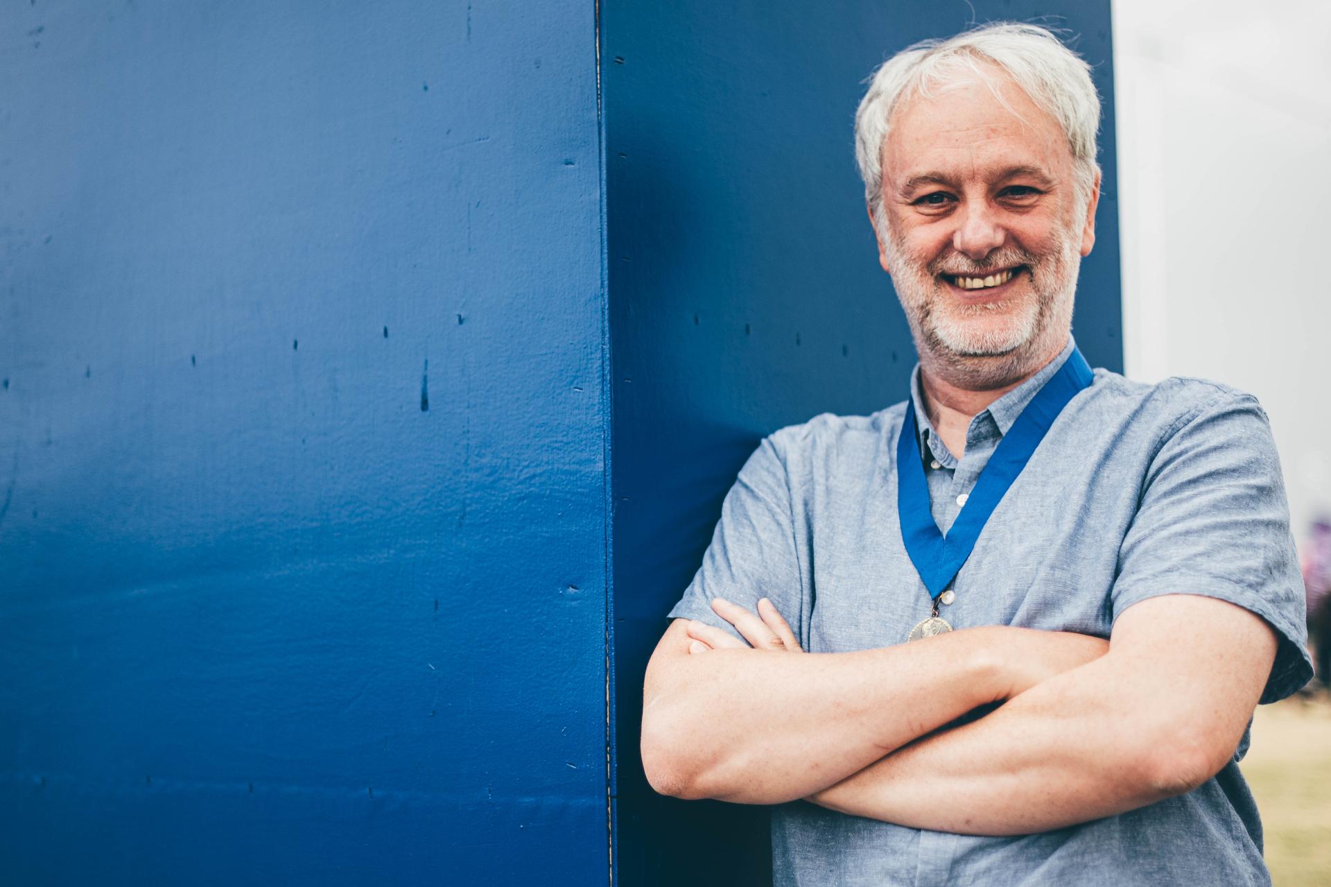 Person with crossed arms wearing a gray shirt and blue lanyard, standing next to a blue wall.