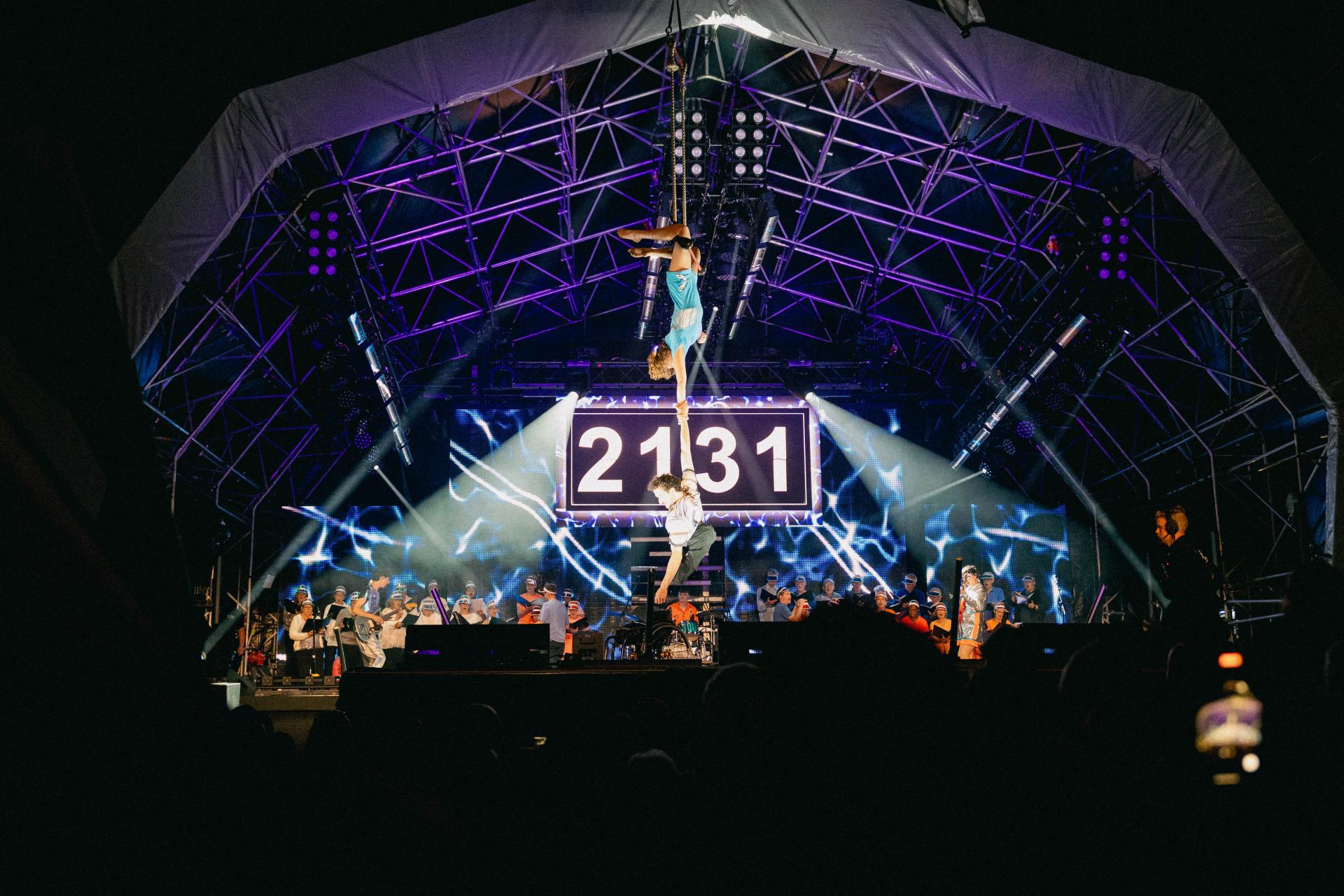 Stage performance with acrobats suspended in the air, large digital display showing '2131', musicians and performers on stage, under bright lights and metal framework