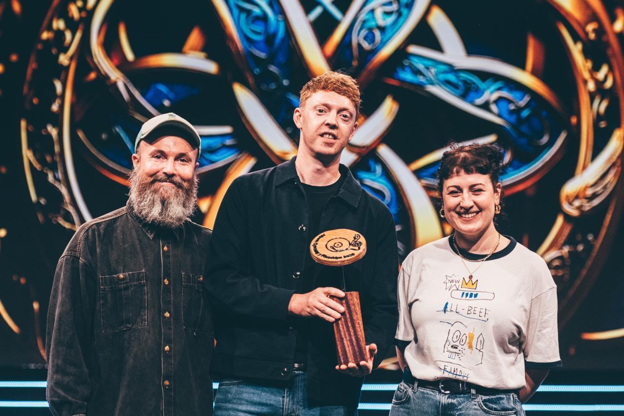 Three individuals standing in front of a colorful decorative background, with the central figure holding a wooden award or trophy;