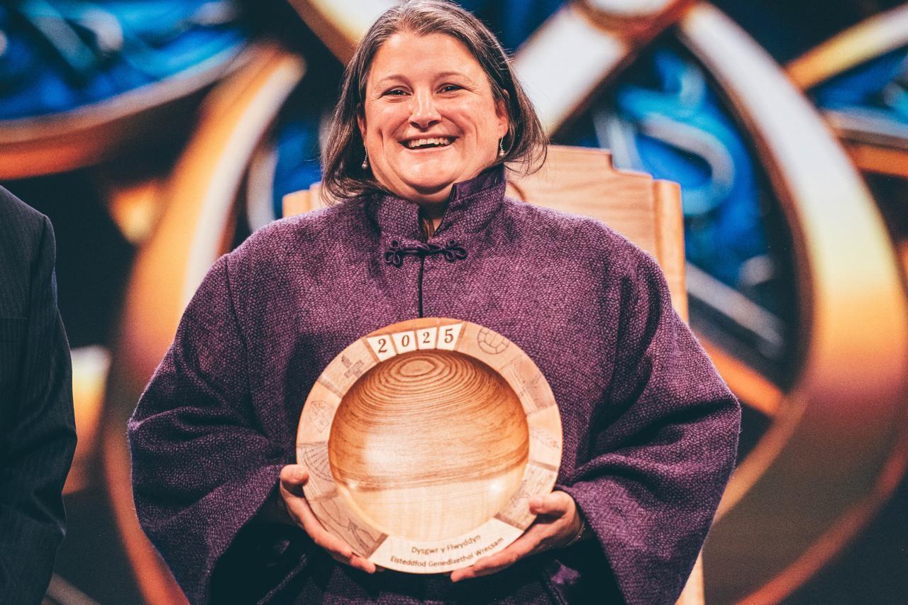 Person in a purple garment holding a wooden award engraved with '2025' and the words, Dysgwr y Flwyddyn