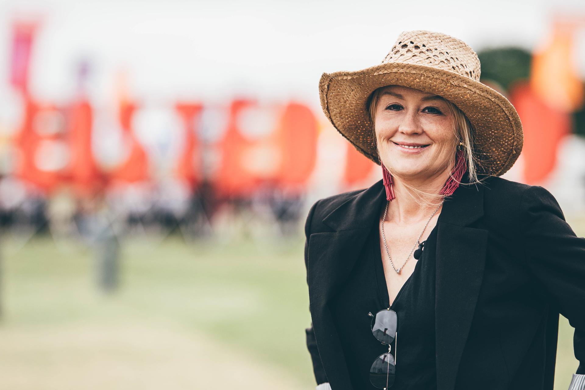 Person wearing a straw hat, black blazer, and sunglasses hanging from the blazer at an outdoor event with red banners and blurred crowd in the background