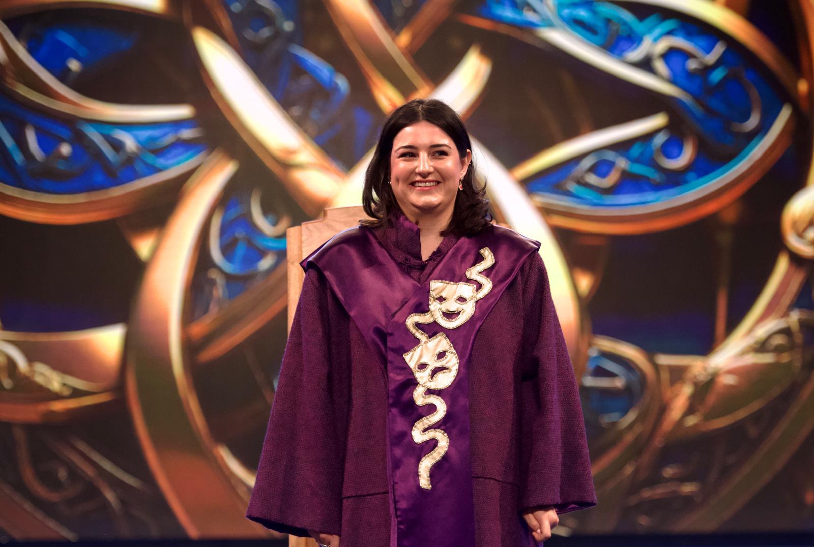 Person in a purple robe with theatrical masks and snake design, standing before an ornate, colourful patterned background.