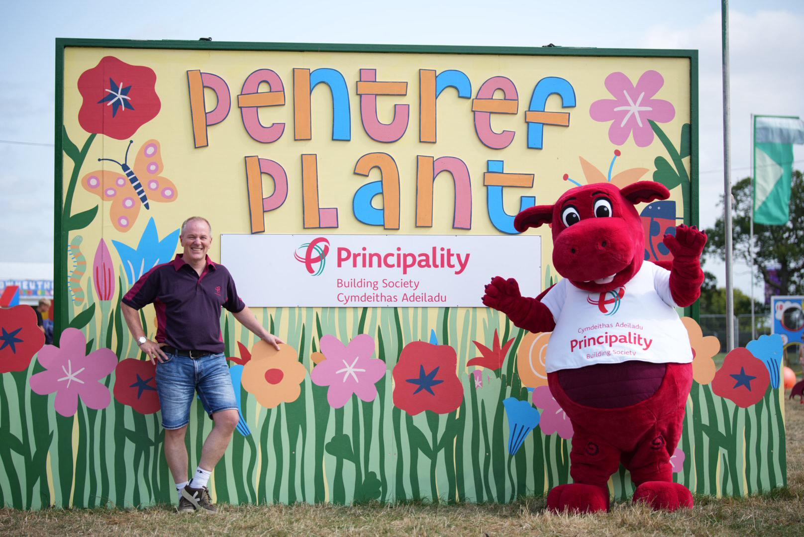 Colorful 'Pentref Plant' sign with flowers and a butterfly, featuring the Principality Building Society logo; a person stands on the left and a red dragon mascot wearing a 'Principality' shirt on the right