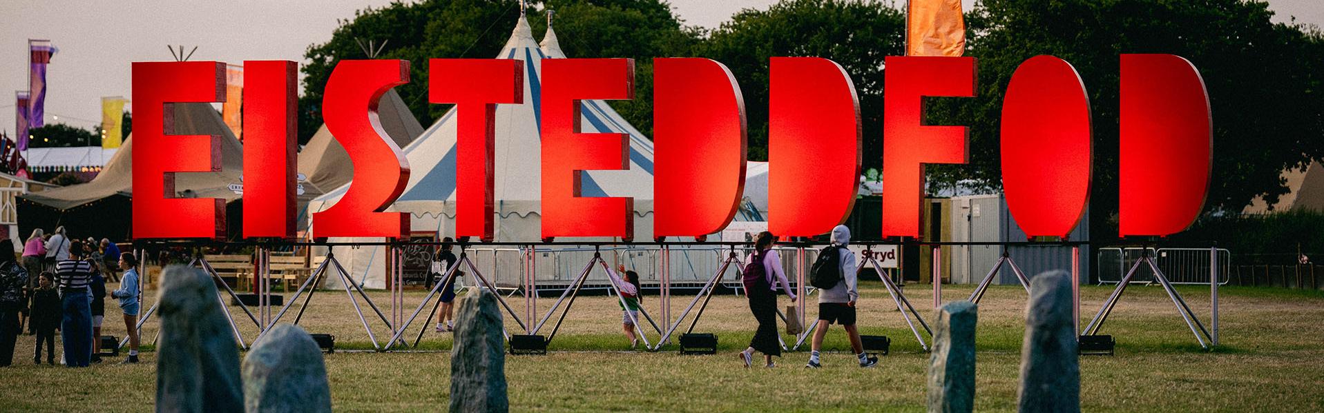 Outdoor event with large red 'EISTEDDFOD' letters, people walking among tents and trees, and a circle of standing stones in the foreground