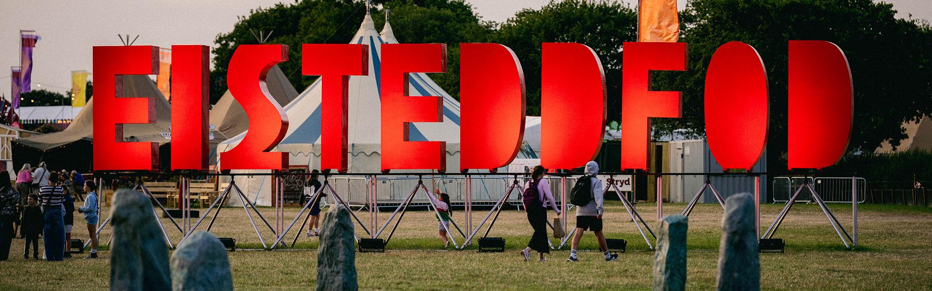 Outdoor event with large red 'EISTEDDFOD' letters, people walking among tents and trees, and a circle of standing stones in the foreground